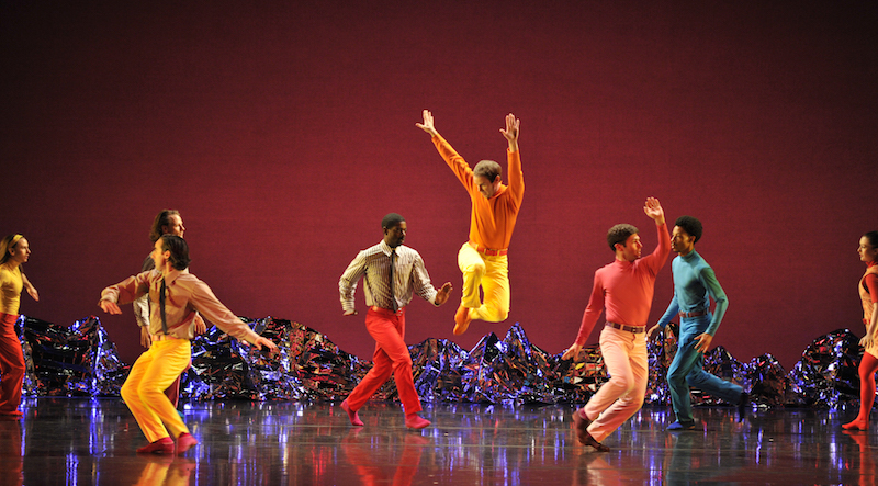 A dancer in orange and yellow leaps high into the air while other dancers, some in turtlenecks and others in ties, move around the solo jumper.